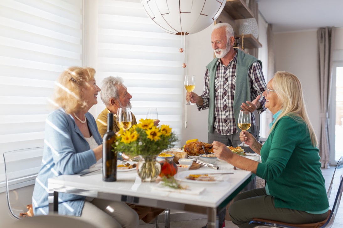 Two senior couples gathered around table, celebrating Thanksgiving, having dinner together at home and raising glasses of wine, making a toast, host welcoming guests