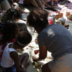Um grupo de pessoas está reunido em um piquenique ao ar livre, sentado sobre uma toalha xadrez. Algumas crianças estão presentes, e uma mulher está agachada, cortando frutas, possivelmente para compartilhar. As pessoas ao redor parecem estar envolvidas em atividades relaxantes, como comer e socializar. O ambiente é iluminado pela luz natural, sugerindo um dia ensolarado. Há cestas e frutas visíveis ao fundo, criando uma atmosfera acolhedora e comunitária.
