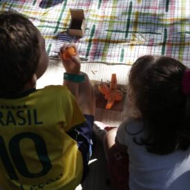 Duas crianças estão sentadas em um chão coberto por uma toalha de piquenique. A criança à esquerda, vestindo uma camiseta amarela com a palavra "BRASIL" e o número 10, está segurando um brinquedo laranja. A criança à direita, com um laço rosa no cabelo, observa e parece estar brincando com um avião laranja. Ao fundo, há uma caixa de papel e a luz do sol ilumina a cena, criando um ambiente alegre e descontraído.