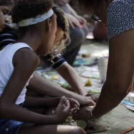 Um grupo de crianças e uma adulta estão sentados em um ambiente ao ar livre, participando de uma atividade manual. As crianças estão concentradas, com as mãos unidas, enquanto a adulta, que está à direita, está ajudando-as. O chão é coberto por um tapete ou tecido colorido, e há diversos materiais ao redor, possivelmente para artesanato. As crianças têm expressões de curiosidade e envolvimento na atividade. A cena transmite um sentimento de aprendizado e colaboração.
