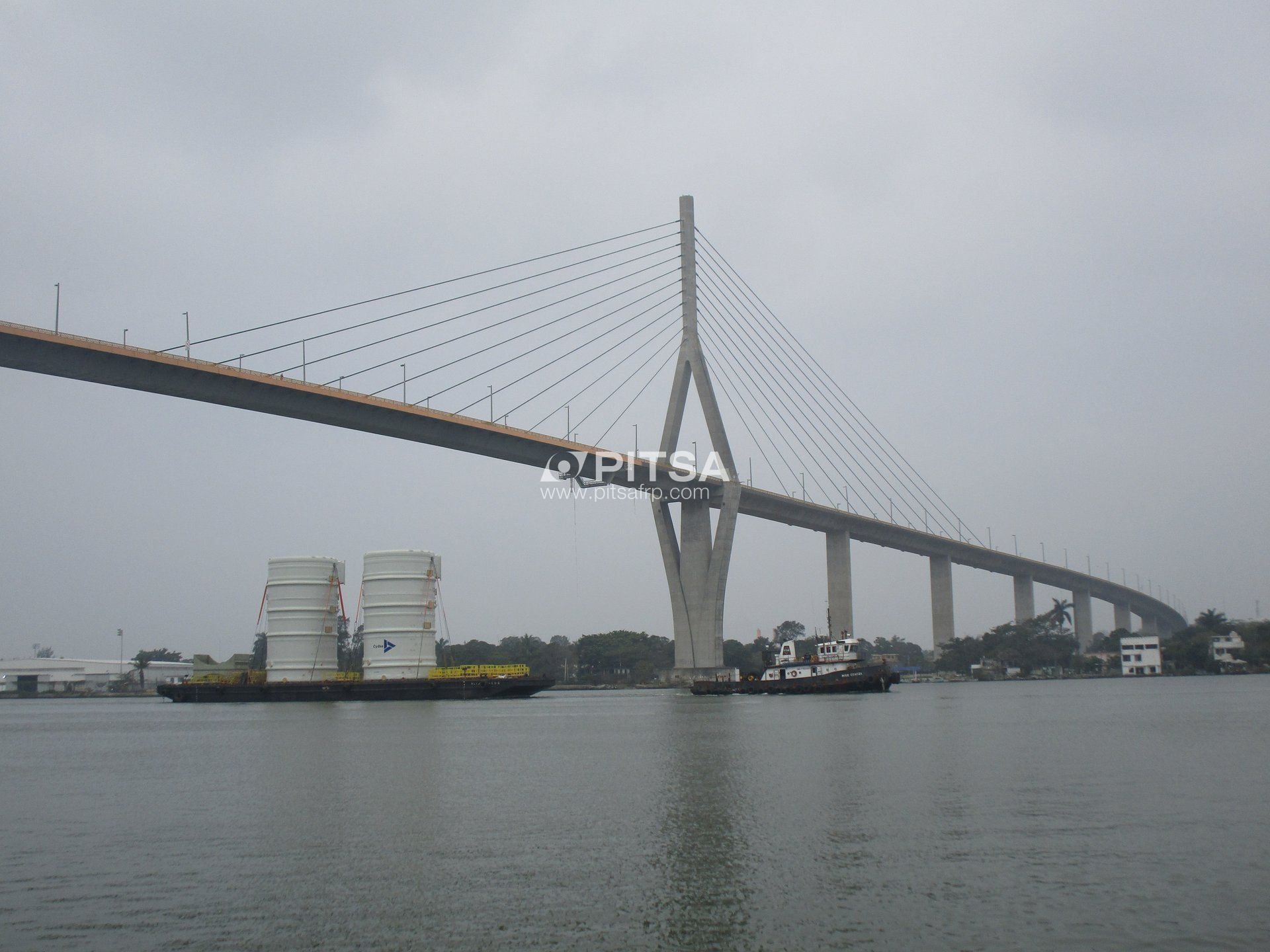 Two large white fiberglass reinforced plastic (FRP) tanks on a barge being transported under a cable-stayed bridge.