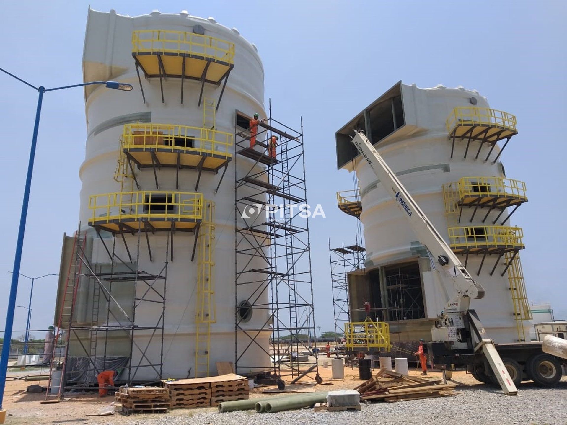 Two large white FRP tanks with yellow gratings and stairs installed at a construction site, featuring scaffolding and crane equipment.
