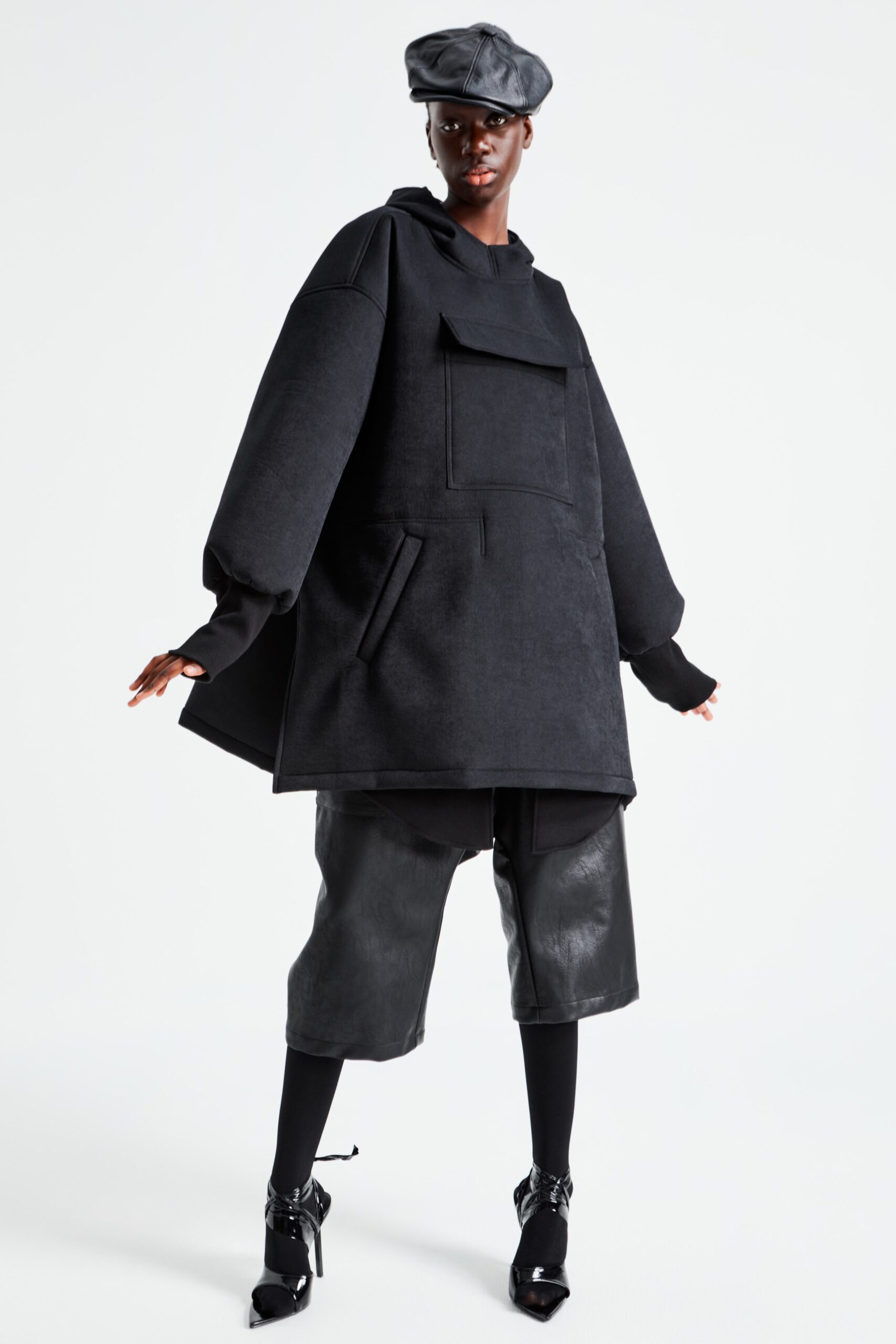 An oversized black hoodie features a unique pocket detail and a relaxed silhouette, creating a contemporary streetwear vibe. Paired with sleek leather shorts, this ensemble balances casual comfort with a bold, structured aesthetic.