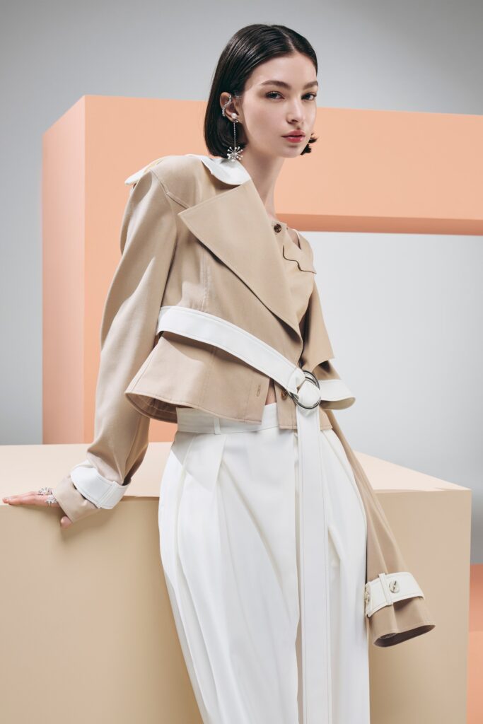 A fitted beige jacket with long sleeves and a belted design, paired with white pants.