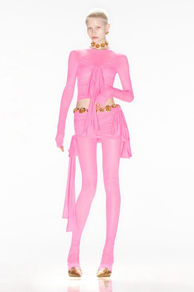 A fitted pink silk dress with long sleeves, a V-neckline, and asymmetric ruched details paired with fitted pants.