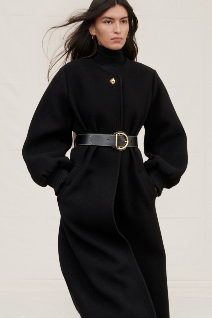 A relaxed black wool coat with long sleeves, high neck, and a belted detail.