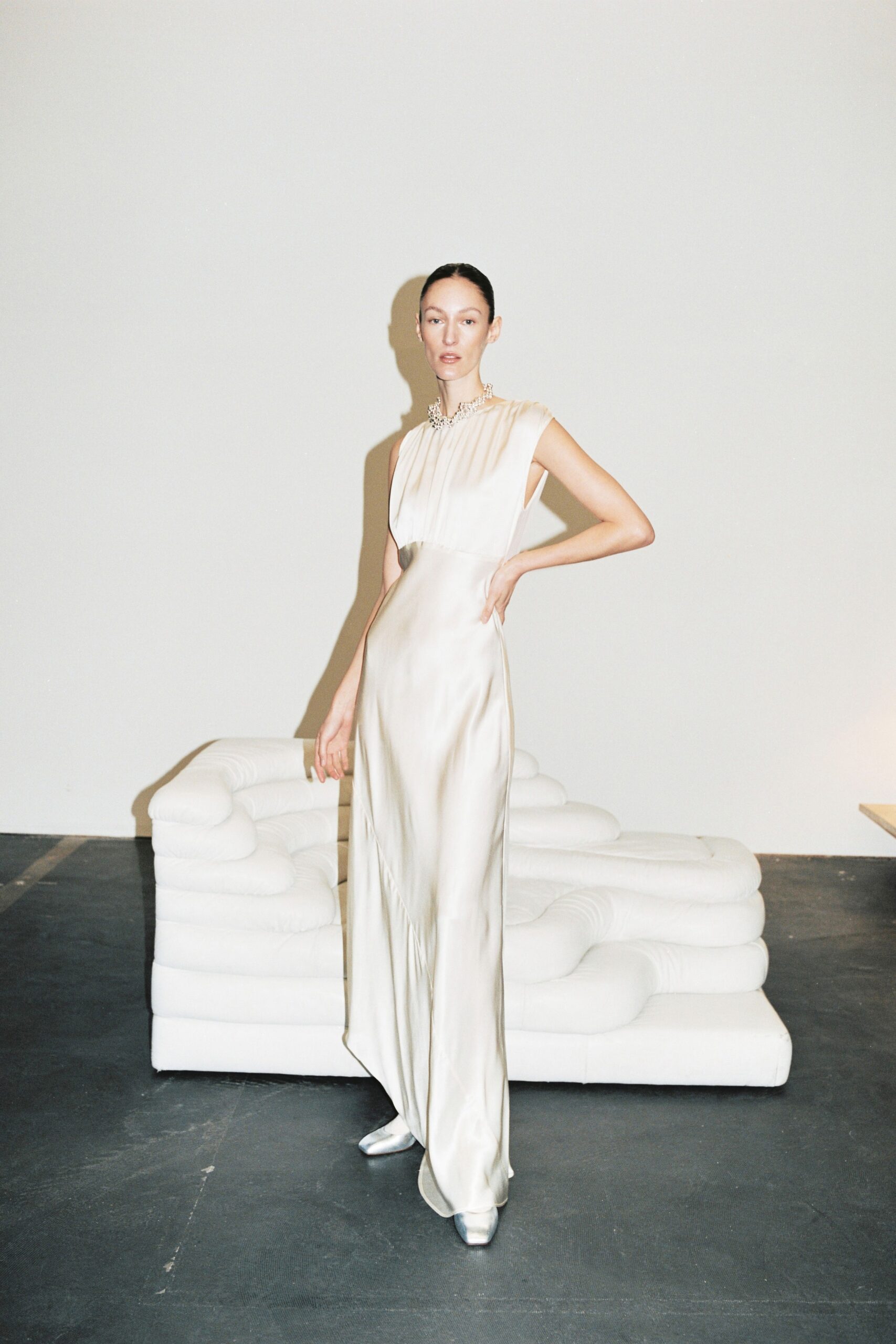 A fitted cream silk sleeveless gown with a high neckline and pleated detailing.