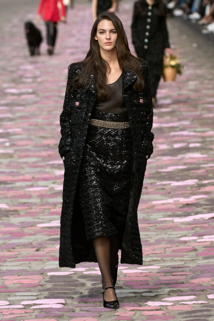 A fitted black sequined dress with a V-neckline and layered pencil silhouette, topped with a long-sleeve coat.