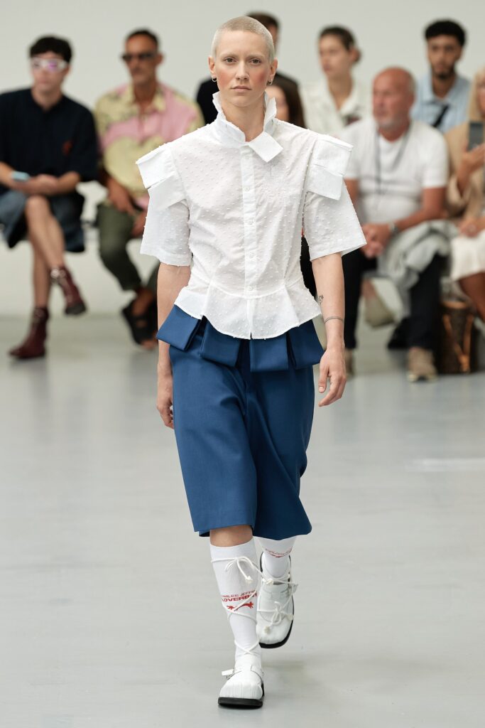 A fitted white cotton top with short sleeves and pleated blue shorts featuring an asymmetric design.