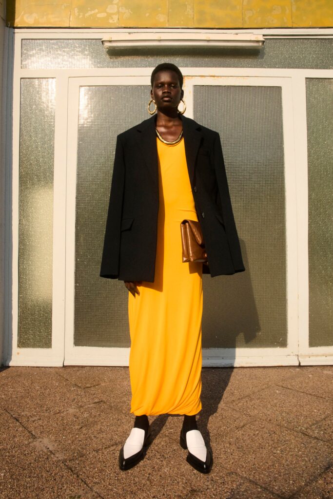 A fitted yellow sleeveless dress paired with a black jacket, featuring a draped design.