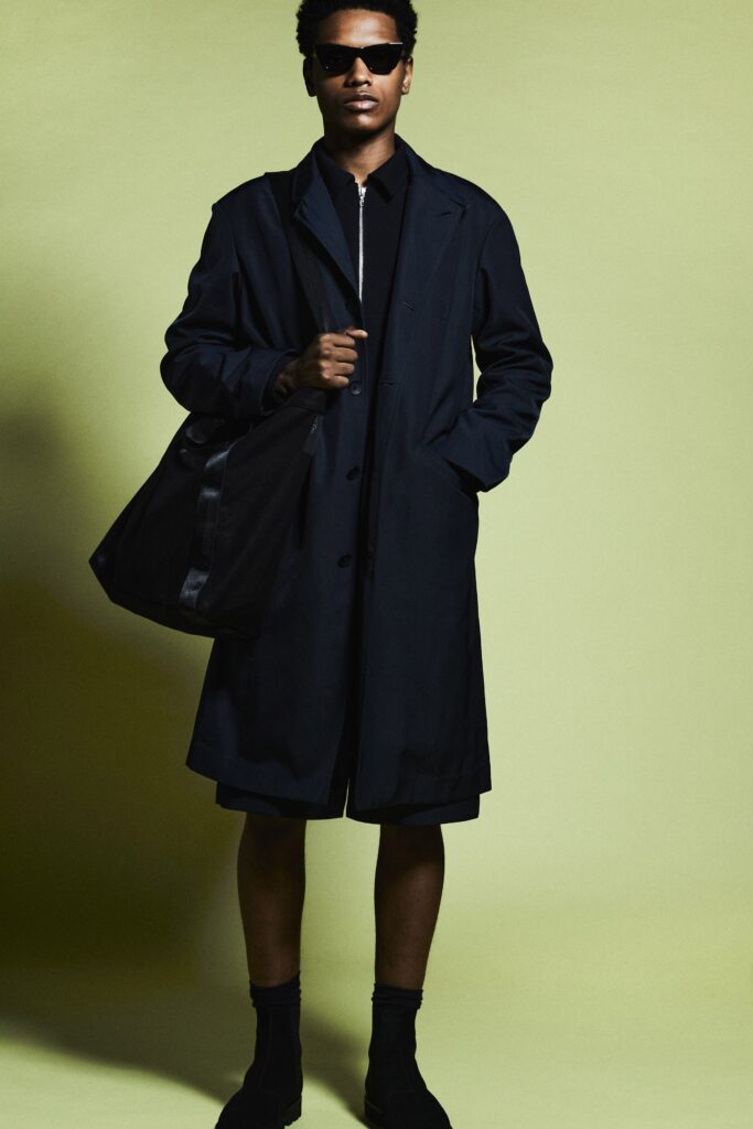 A relaxed black coat paired with collared long-sleeve shirt and black shorts.