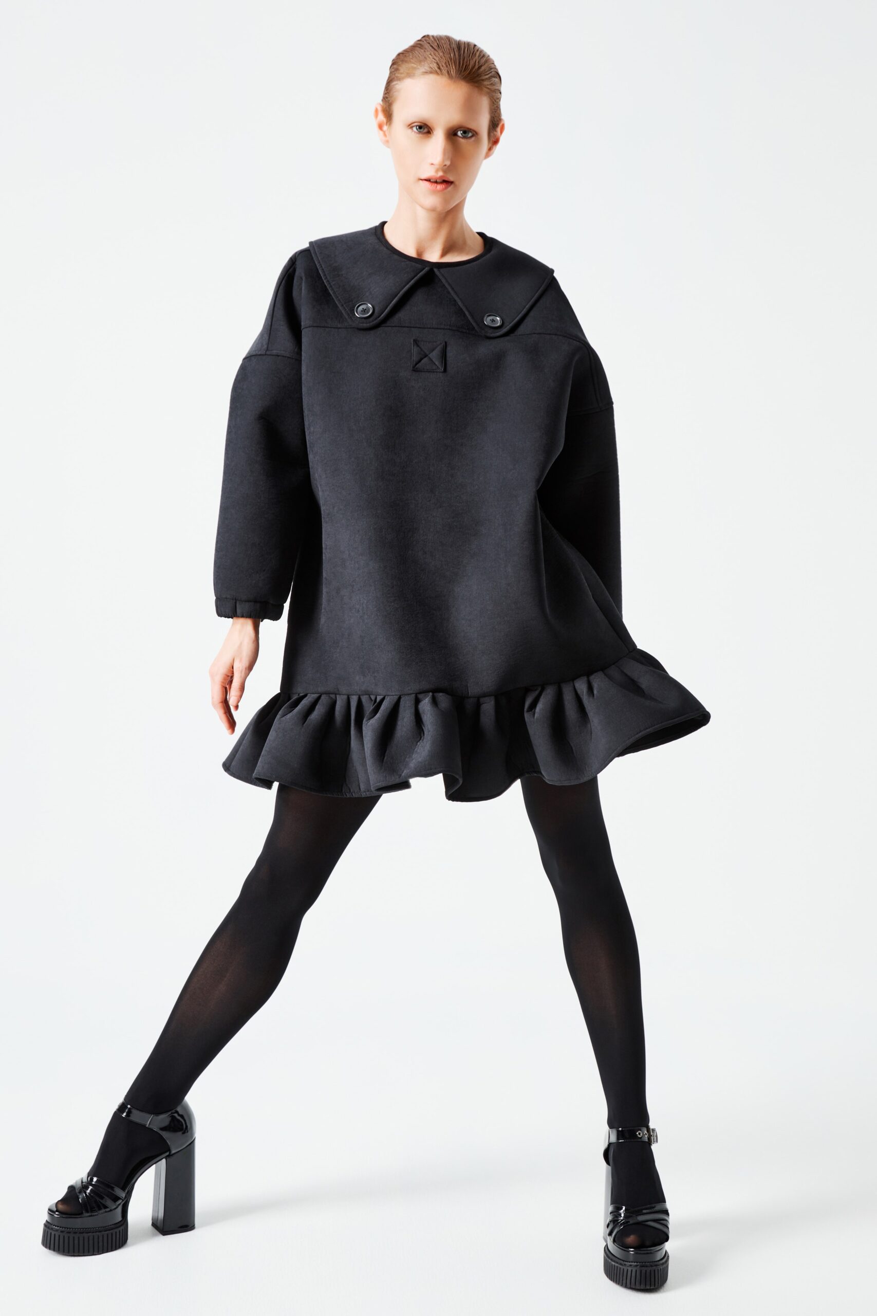 This relaxed dress features a distinctive collar and long sleeves, creating a playful silhouette. The ruffled hem adds movement and dimension, enhancing the overall design with a touch of whimsy.