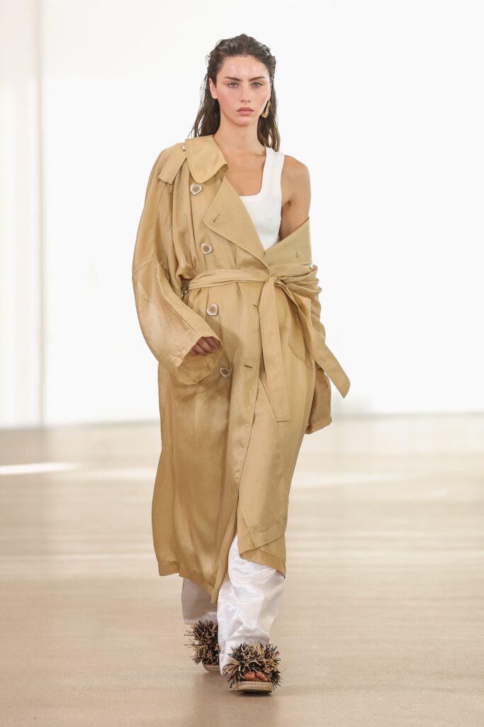 A relaxed beige silk trench coat paired with white cotton pants, featuring a belted design.