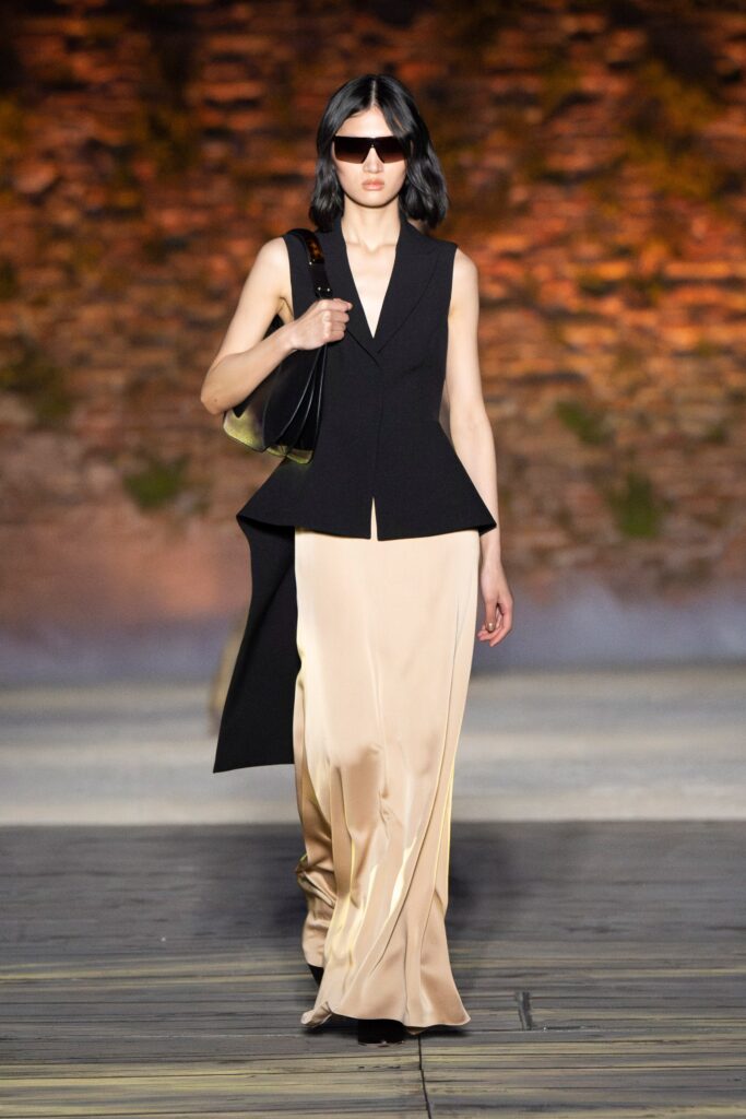 A fitted black sleeveless jacket worn over a beige silk dress with an asymmetric design.