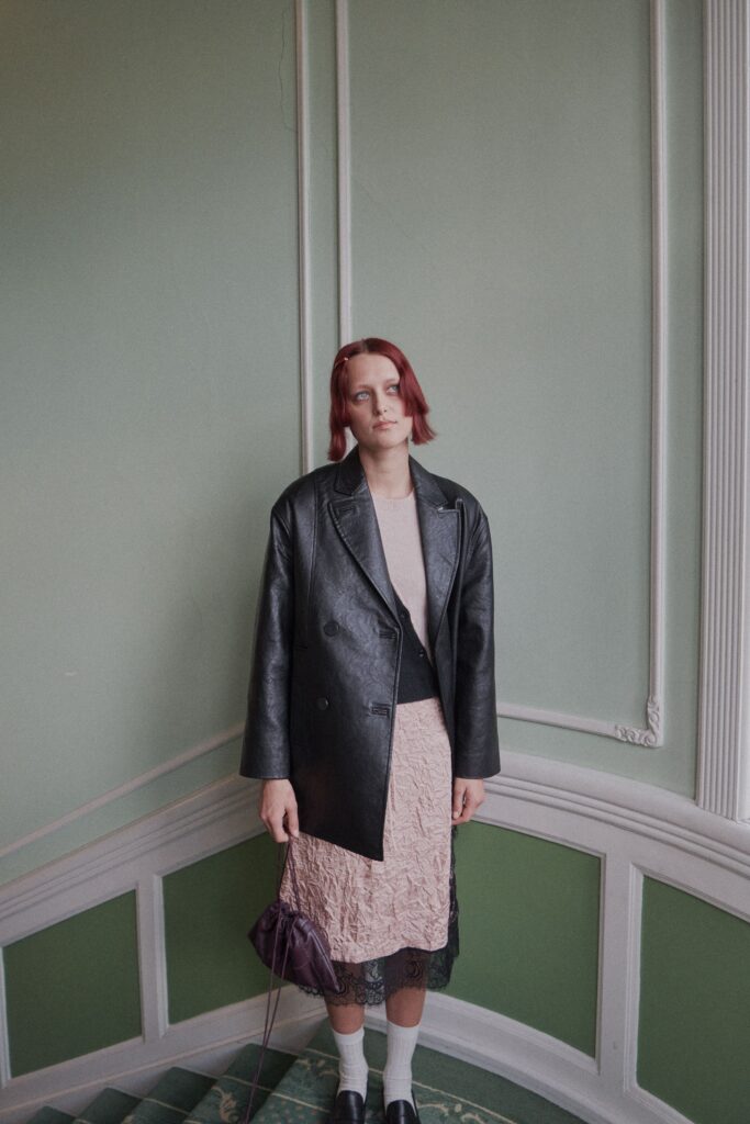 This ensemble features a fitted black leather jacket layered over a pink lace dress, creating a striking contrast in textures. The asymmetric silhouette and V-neckline of the dress add a contemporary edge, while the long sleeves of the jacket provide a structured balance to the overall look.