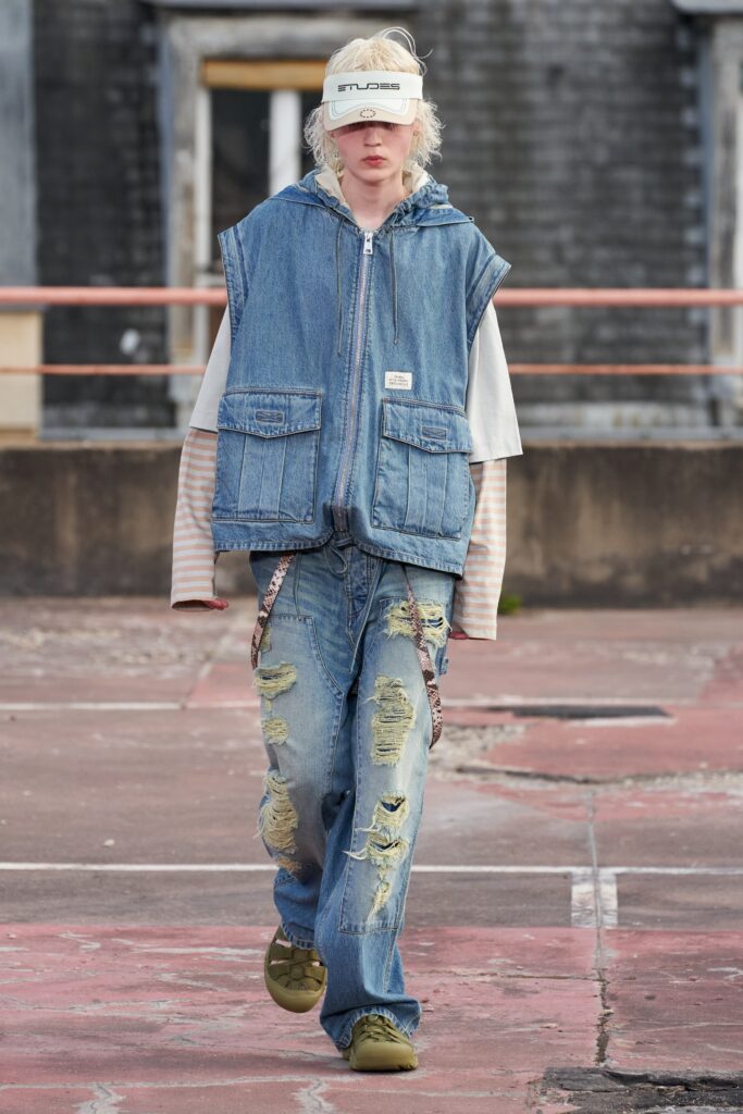 A relaxed blue denim vest worn over a long-sleeve shirt paired with distressed denim pants.