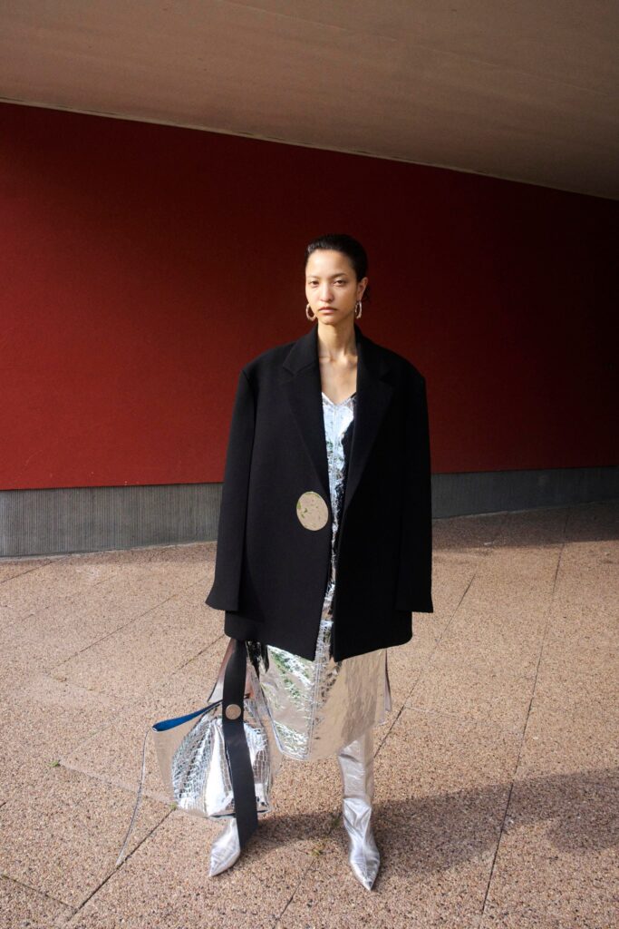 An oversized black coat layered over a silver dress with a V-neckline and asymmetric design.