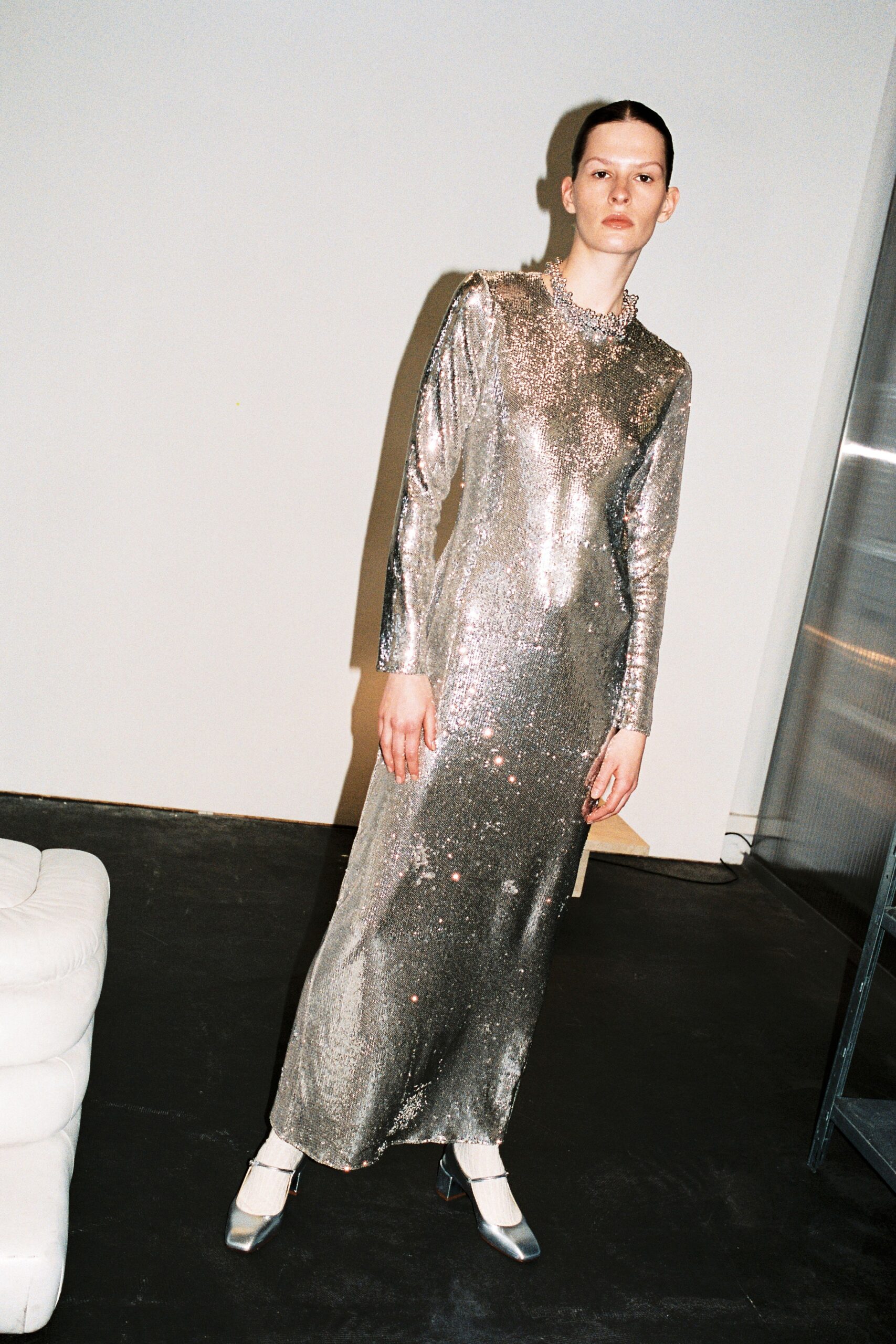 A fitted silver metallic dress with long sleeves and a high neckline featuring a slit.