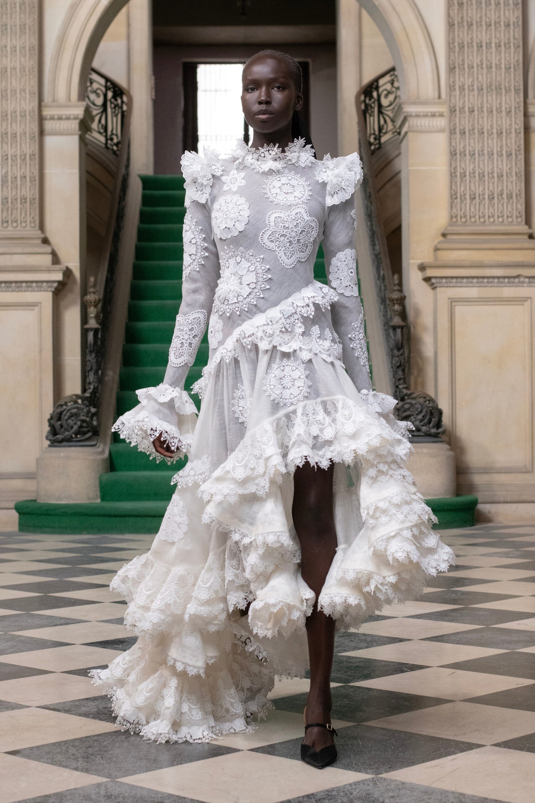 A fitted white lace dress with long sleeves, high neckline, and ruffled asymmetric hem.