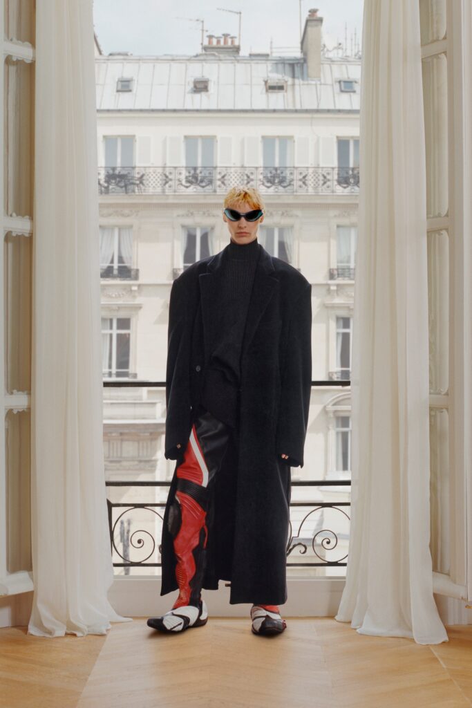 An oversized black coat paired with flared pants featuring red accents and a high-neck design.