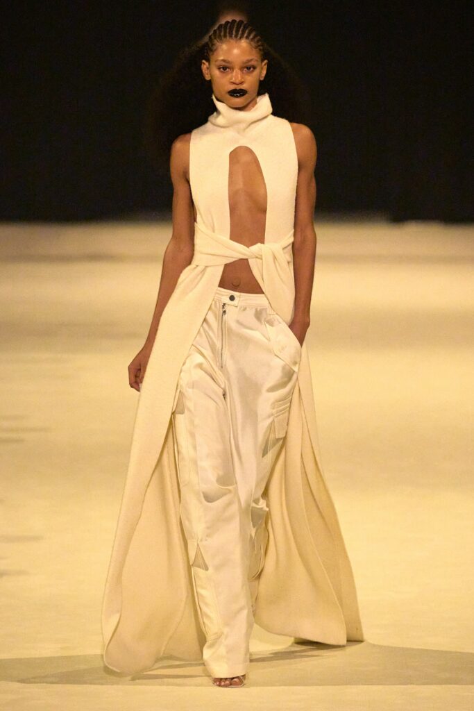 A fitted white sleeveless dress with a V-neckline and asymmetric design paired with pants.