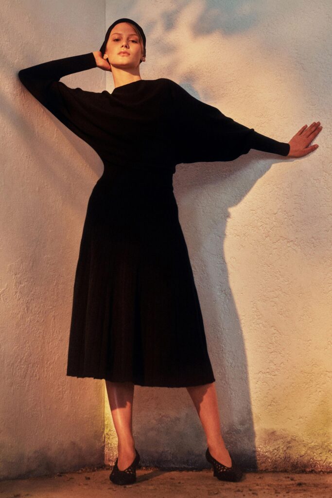 A fitted black knit dress with long sleeves, high neckline, and pleated design.