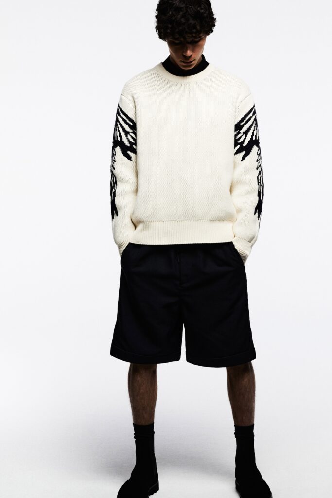 A relaxed white sweater with printed details paired with black shorts.
