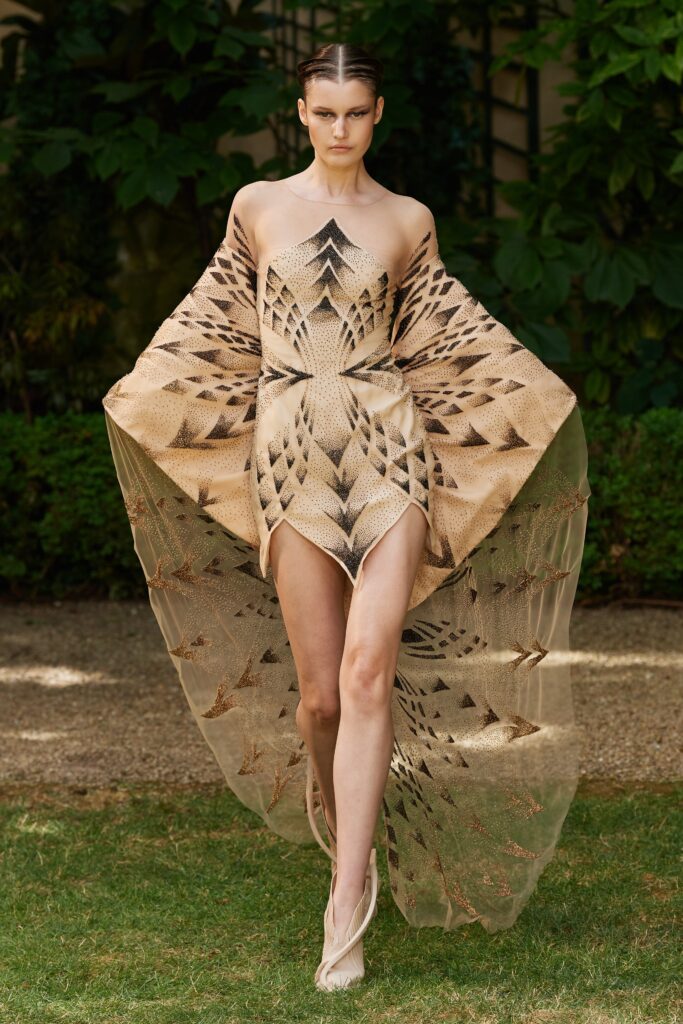 A fitted beige silk dress with a high-neck and an asymmetric printed cape.