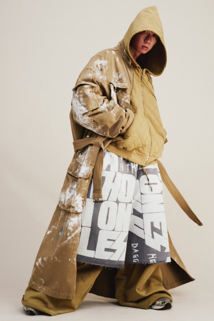 An oversized beige coat with a hood and printed gray pants, creating a layered streetwear look.