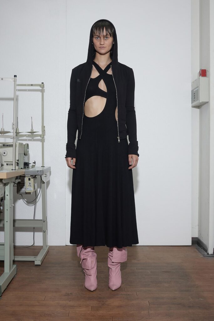 A fitted black sleeveless dress with a hooded jacket and cutout details, paired with pink boots.