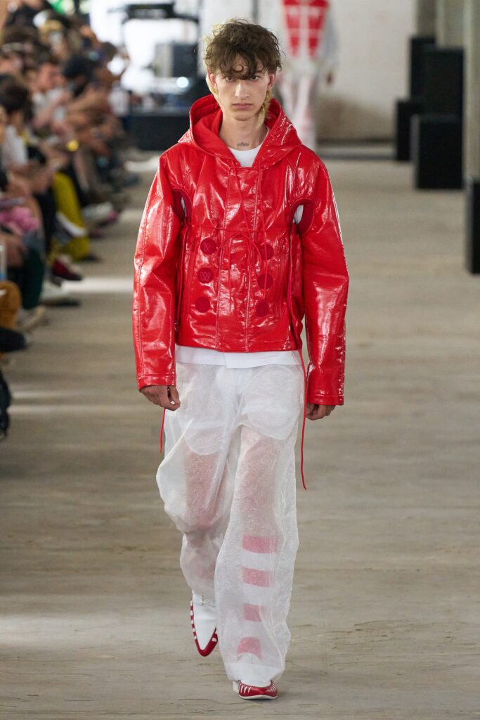 A relaxed red leather hooded jacket paired with white cotton pants featuring a zippered detail.