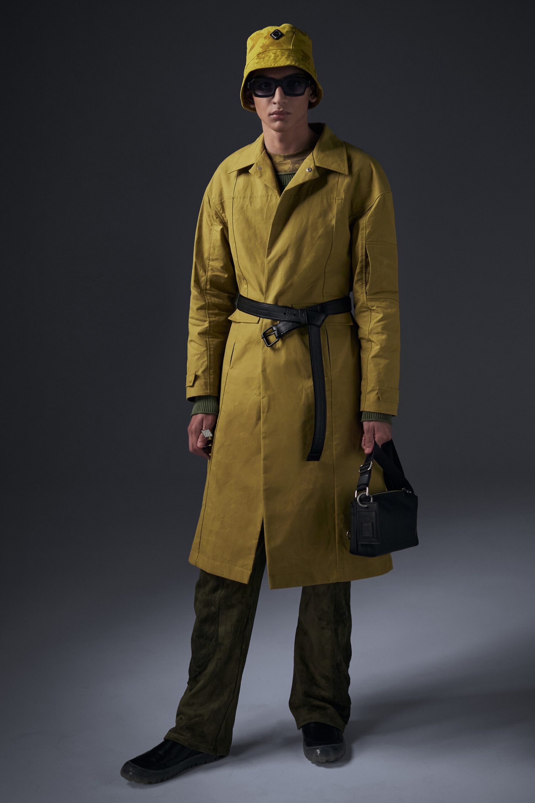 This coat features a relaxed silhouette, accentuated by a belted waist that adds structure while maintaining a laid-back vibe. The vibrant yellow hue contrasts beautifully with the earthy green pants, creating a striking visual interplay that commands attention.