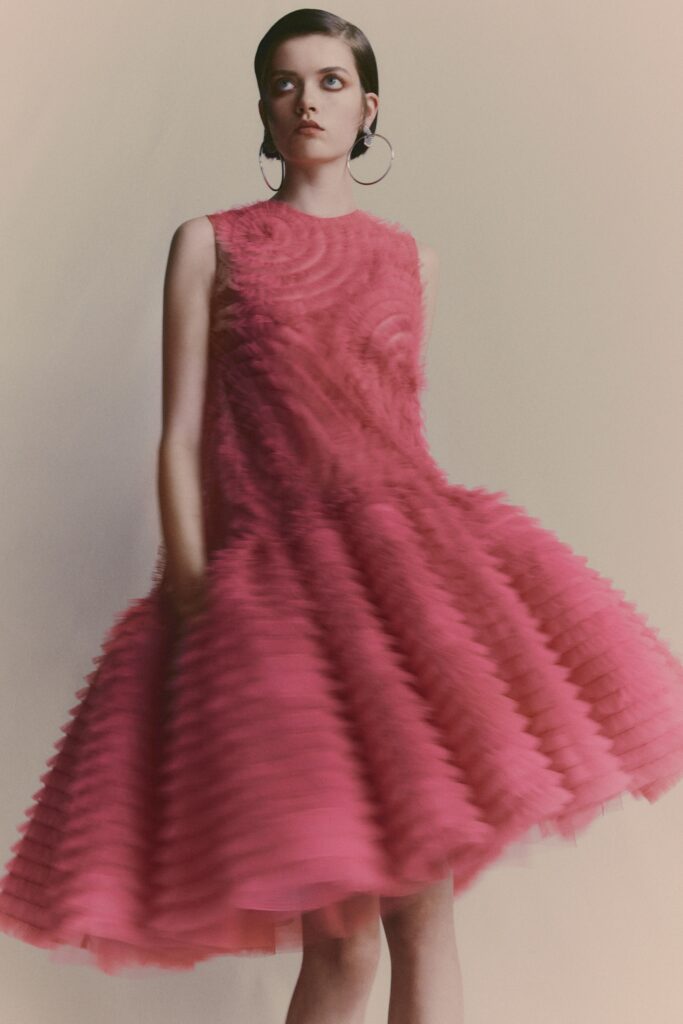 A fitted pink sleeveless dress made of tulle with a high-neck and pleated, tiered design.