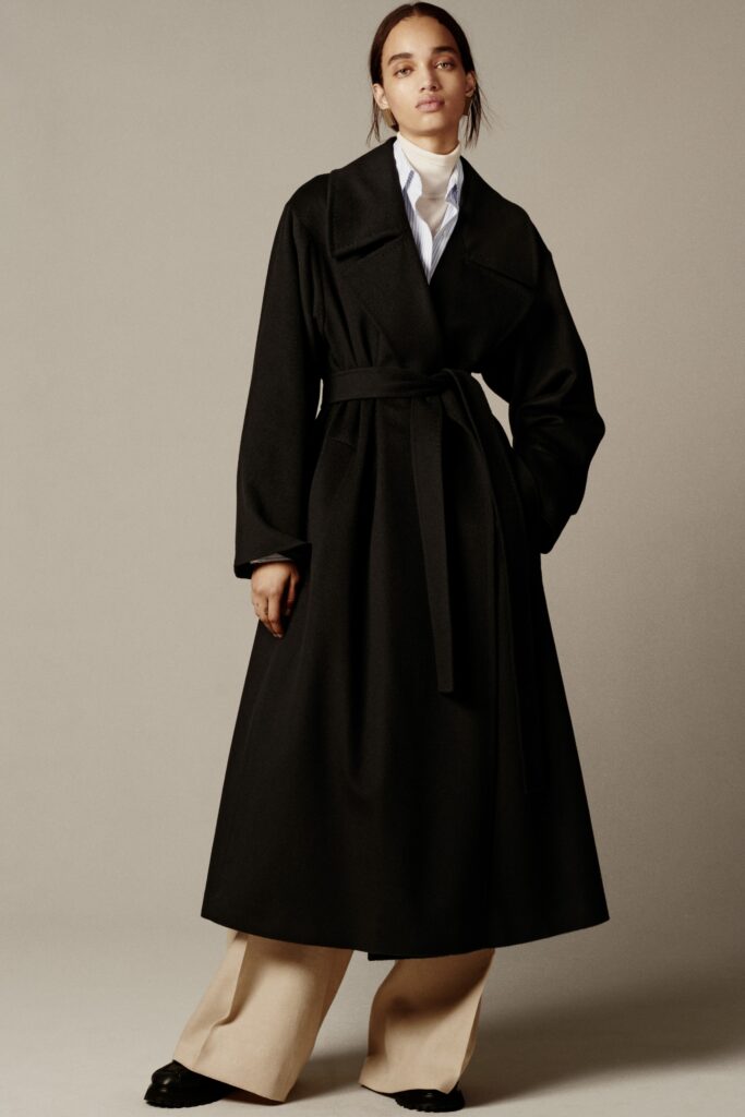 A relaxed black wool coat with a belted waist, paired with beige cotton pants.
