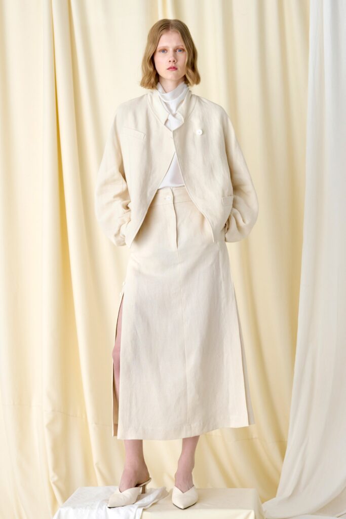 A fitted beige cotton jacket with long sleeves and a collared neckline paired with a slit skirt.