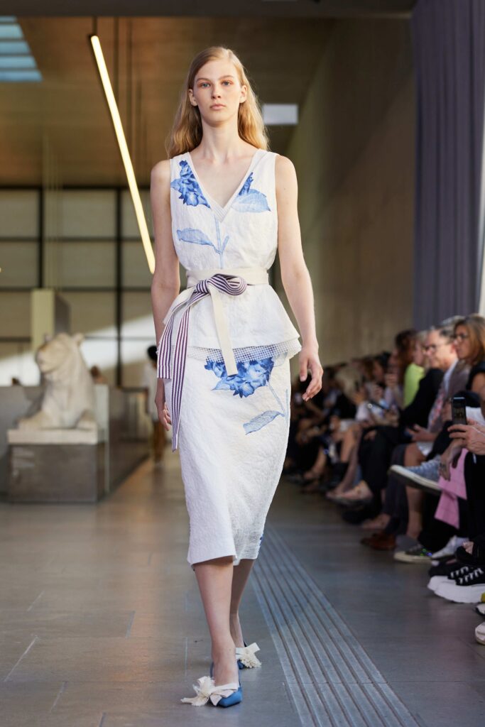 A fitted white cotton sleeveless dress adorned with blue floral patterns and a tied waist detail.