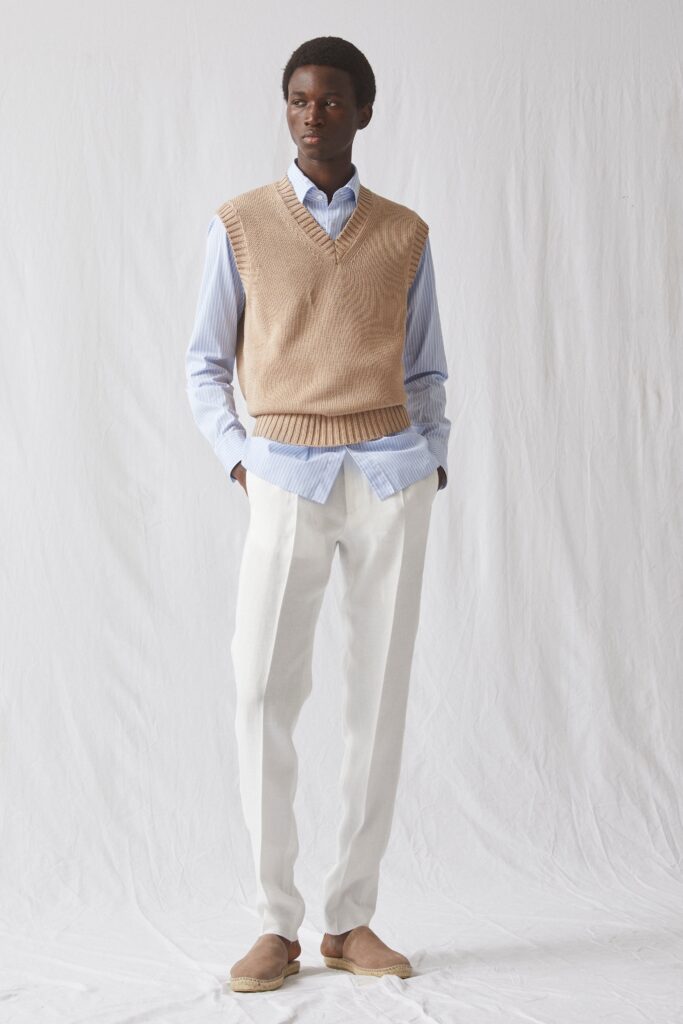 A relaxed beige vest layered over a blue striped long-sleeve shirt paired with white pleated pants.