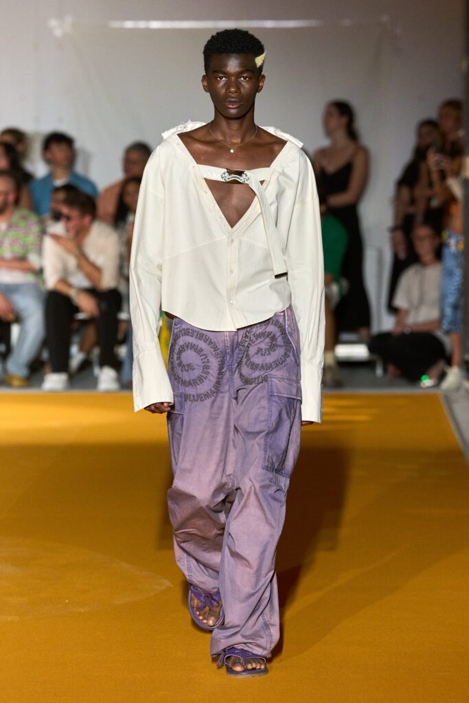 A relaxed white long-sleeve shirt with an asymmetric design paired with embroidered purple pants.