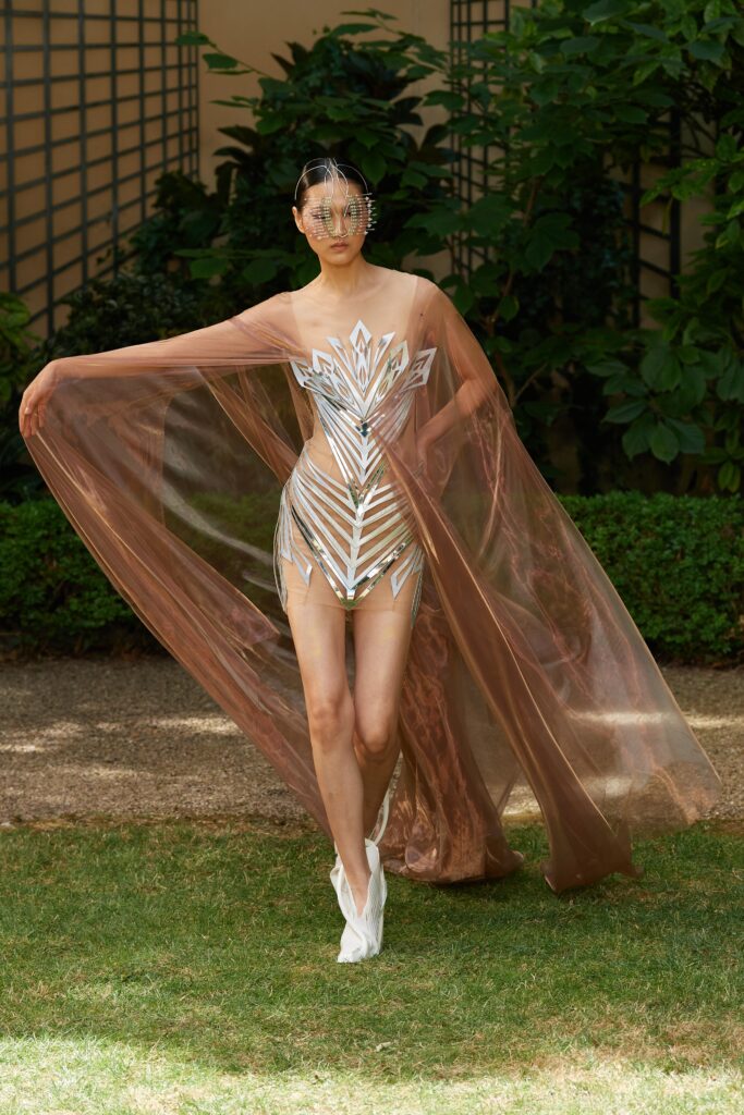 A fitted brown sheer sleeveless dress with a V-neckline and layered asymmetric cape.