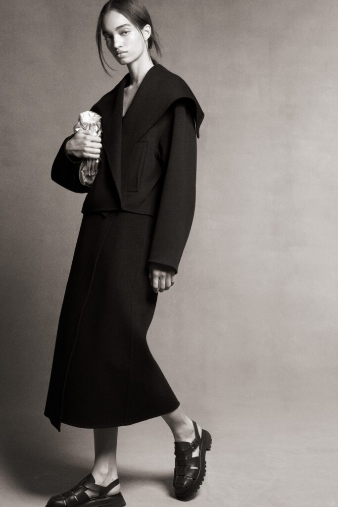 A fitted black wool dress with a long-sleeve collared jacket featuring an asymmetric layered design.