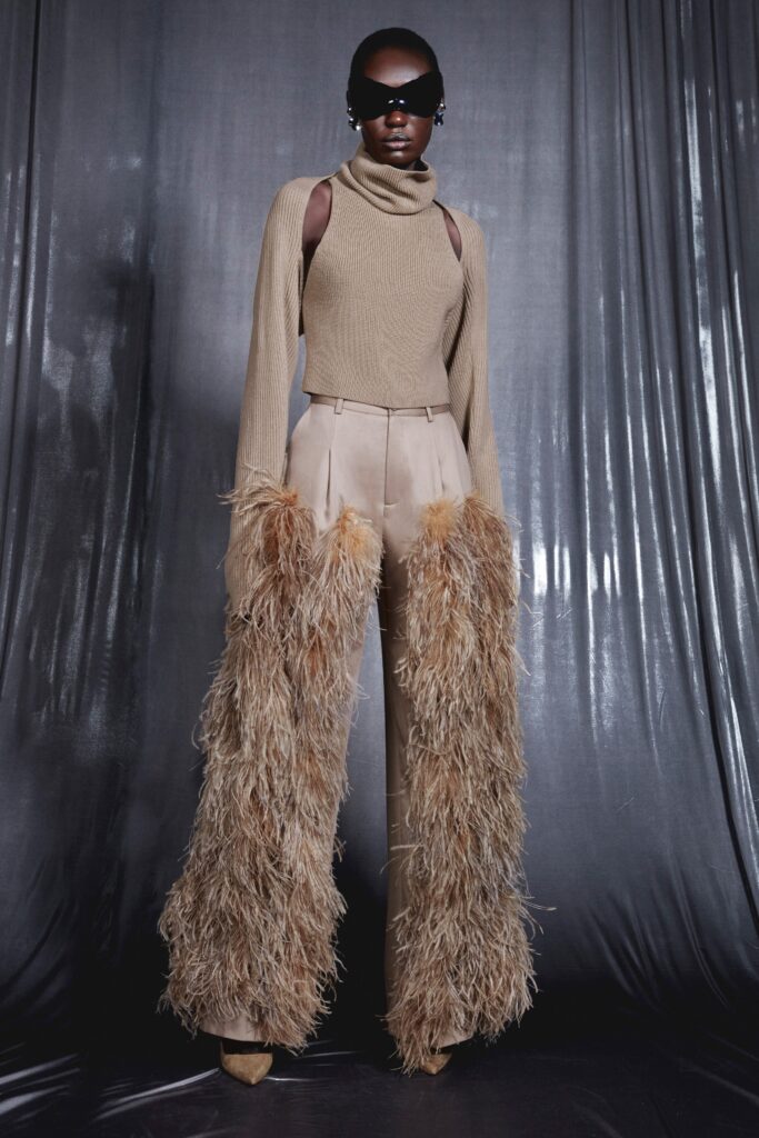 A relaxed beige wool top with long sleeves and a high neck, paired with fringed brown pants.