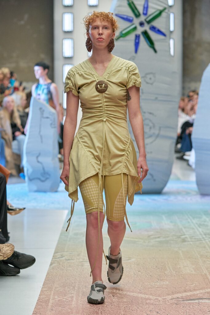 A fitted beige dress with short sleeves and an asymmetric draped design paired with shorts.