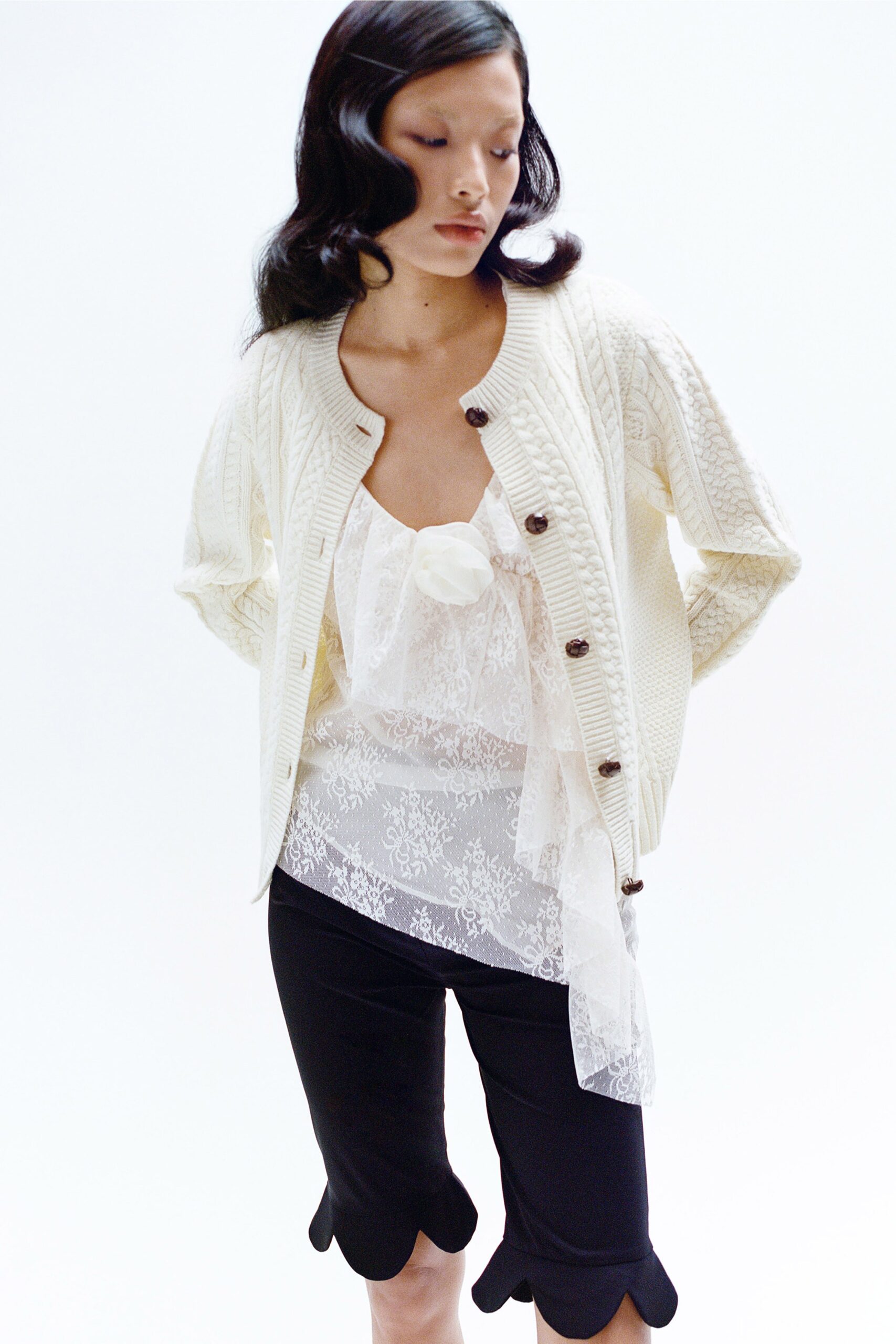 A fitted white lace top layered under a long-sleeve cardigan with black shorts.