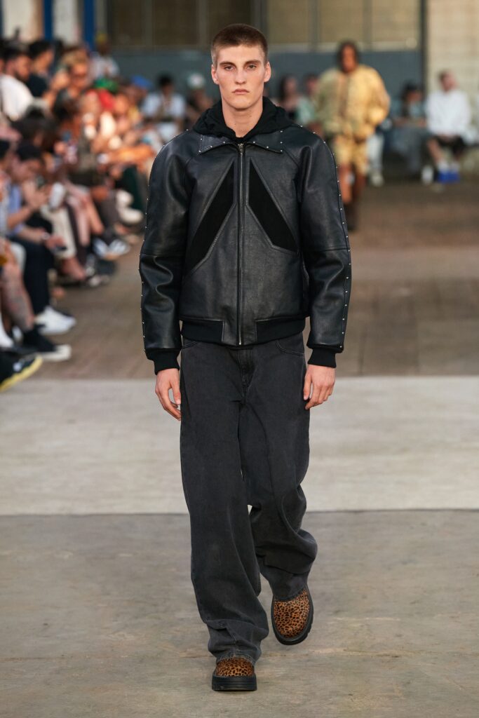 A relaxed black leather jacket with a hood paired with gray denim pants.