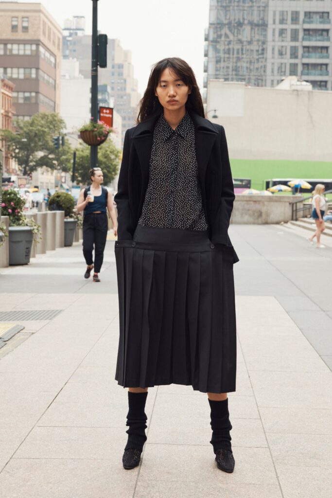 A fitted black coat paired with a pleated black skirt, featuring a collared long-sleeve shirt.