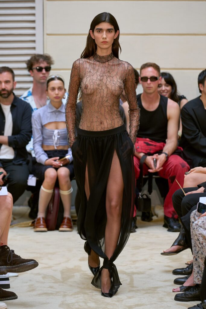 A fitted black sheer dress with long sleeves, high neckline, and a layered silhouette featuring a slit.