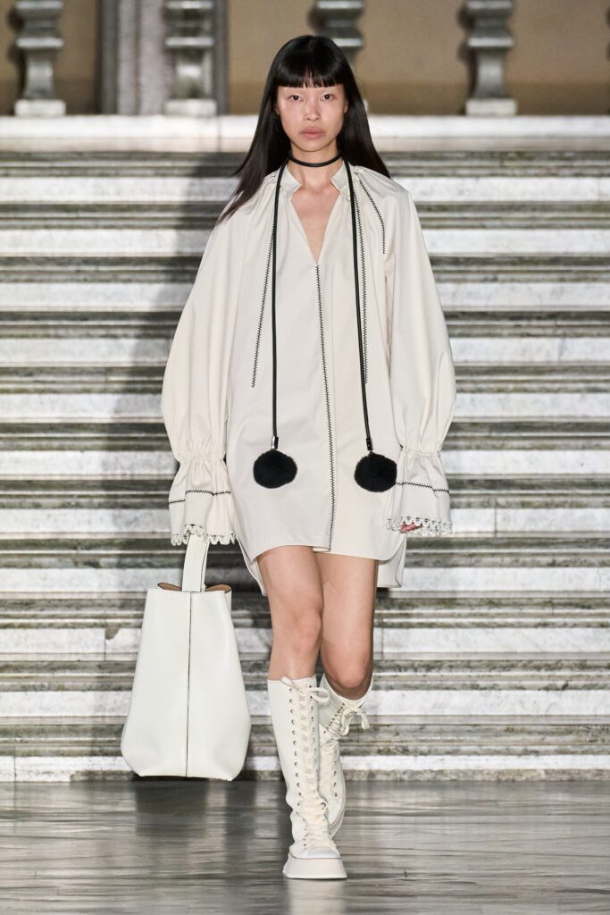 A relaxed white cotton dress with long sleeves and a V-neckline featuring pleated details.
