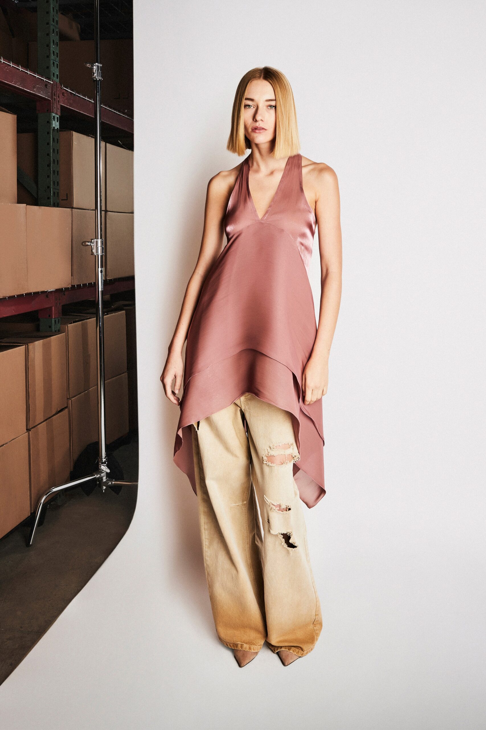 A relaxed pink silk sleeveless top with a V-neck and asymmetric design paired with distressed beige denim pants.