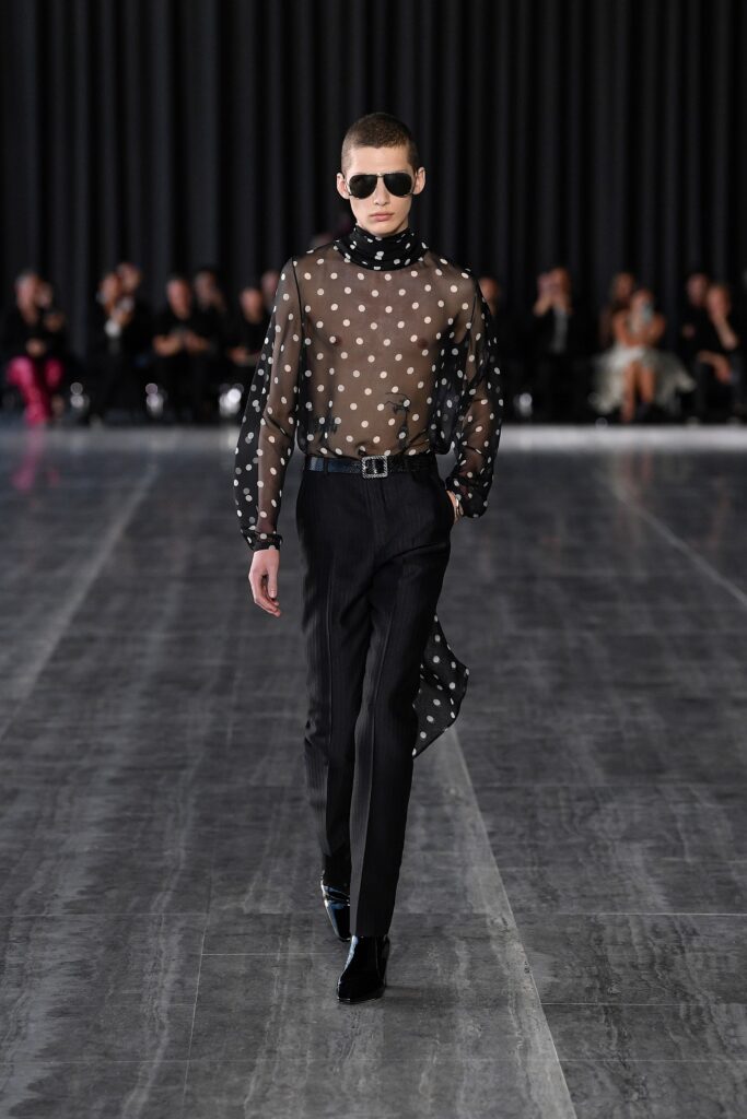 A fitted black sheer polka-dot shirt with long sleeves and asymmetric design paired with fitted pants.