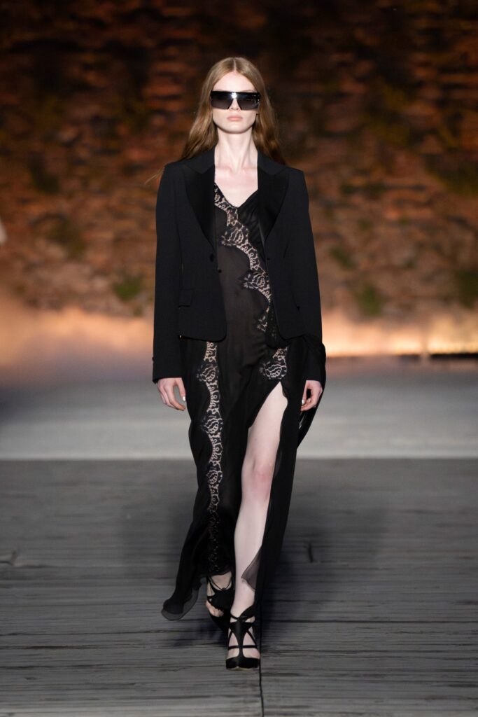 A fitted black sheer dress with a V-neckline and slit, paired with a black jacket.